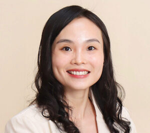 Headshot of a smiling person with long dark hair, wearing a light-colored blazer, set against a plain background.