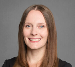 Van Nostrand, Caneel Eva_final Headshot of a person with long hair, smiling, against a neutral gray background, wearing a dark outfit.
