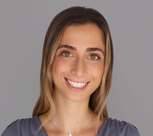 Headshot of a smiling person with long hair wearing a gray top and necklace, against a plain background.