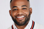 Headshot of a smiling individual with a short beard and closely shaved hairstyle, wearing a white shirt and sweater with a striped collar.