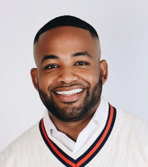 Walker, James Lorenzo III_final Headshot of a smiling individual with a short beard and closely shaved hairstyle, wearing a white shirt and sweater with a striped collar.