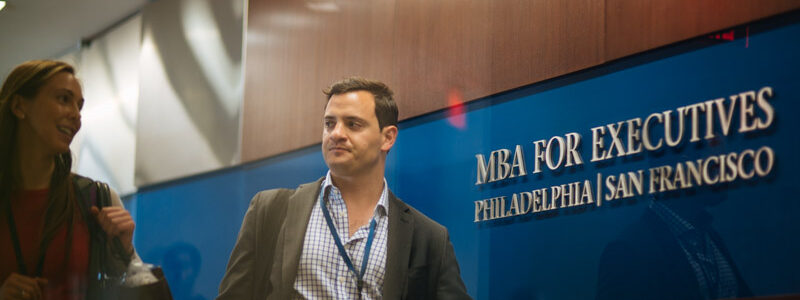 A person walking past a sign for MBA for Executives, Philadelphia | San Francisco, inside a professional setting. Another person is visible nearby.