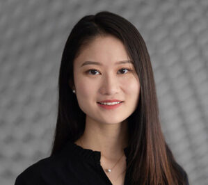 Headshot of a person with long dark hair and a black top, set against a textured gray background.