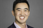 This is a headshot of a smiling person wearing a dark blazer and light-colored shirt against a plain background.